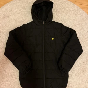 Svart pufferjacka från Lyle & Scott - Säljer en svart pufferjacka från Lyle & Scott med huva och dragkedja. Jackan har ett gult emblem på bröstet och är tillverkad i polyester. Perfekt för kyliga dagar. Ny pris för en sådan här ca 1200 kr