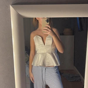 Vit axelbandslös topp - Säljer en stilren vit axelbandslös topp med volangdetalj i midjan. Toppen har en djup v-ringning och är perfekt för en elegant look.