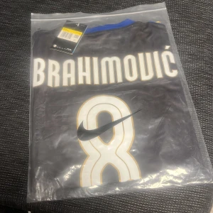 Svart fotbollströja med namntryck - Snygg svart fotbollströja från Nike med namntryck på ryggen. Tröjan har korta ärmar och detaljer i blått och guld. Perfekt för fotbollsfans som vill visa sitt stöd. Zlatan Ibrahimovic 2008 