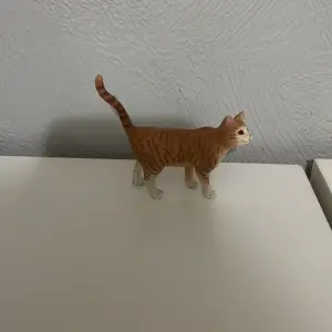 En liten leksakskatt i plast med orange och vita ränder. Katten står upp med svansen i luften och har detaljerade ansiktsdrag. Perfekt för samlare eller som leksak för barn.