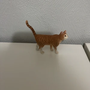 Schleich katt - En liten leksakskatt i plast med orange och vita ränder. Katten står upp med svansen i luften och har detaljerade ansiktsdrag. Perfekt för samlare eller som leksak för barn.