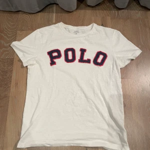 Vit t-shirt från Ralph Lauren - Säljer en vit t-shirt från Ralph Lauren med texten 'POLO' i blått och rött på framsidan. T-shirten är kortärmad och tillverkad i bomull, perfekt för en avslappnad stil. Tröjan har storlek S och modellen är 170cm och 65kg