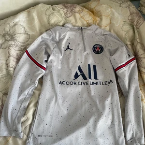 Grå PSG fotbollströja från Jordan - Säljer en grå långärmad PSG fotbollströja från Jordan med svart och rött mönster. Tröjan har en halv dragkedja och klubbens emblem på bröstet. Perfekt för fans av Paris Saint-Germain!