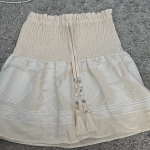Beige kjol med snörning - Charmig beige kjol från Zara med elastisk midja och dekorativ snörning framtill. Kjolen har ett subtilt mönster och är perfekt för varma dagar. Den är helt ny och är i strl XS😊
