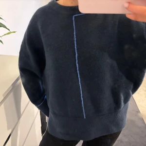 Mörkblå tröja med blå detalj - Säljer en mysig mörkblå tröja med en unik blå linje som detalj framtill. Tröjan har en avslappnad passform och långa ärmar, perfekt för kyligare dagar. Den är enkel men stilren och passar bra till både jeans och leggings.