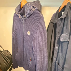 Mörkblå jacka från Moncler - Säljer en snygg mörkblå jacka från Moncler med quiltad design på baksidan och stickade ärmar. Jackan har en huva och dragkedja framtill. Perfekt för kyligare dagar. Skick 10/10, knappt använd. Vid funderingar är det bara att höra av sig!