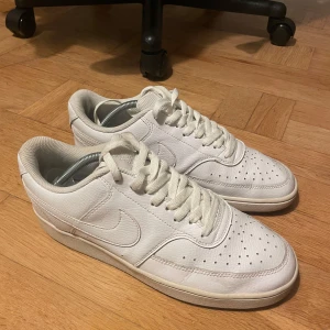 Vita Nike Sneakers storlek 43 - Säljer ett par klassiska vita sneakers från Nike med snörning och perforerade detaljer för extra ventilation. Skorna har en stilren design med den ikoniska Nike-loggan på sidan.