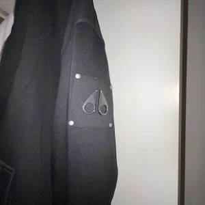 Svart jacka från Moose Knuckles - Säljer en svart jacka från Moose Knuckles med dragkedja och huva. Jackan har en quiltad insida och en snygg logga på ärmen. Perfekt för kyliga dagar med sin varma och stilrena design.
