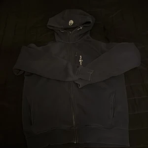 Svart hoodie från Sail Racing - Säljer en svart hoodie från Sail Racing med dragkedja och huva. Perfekt för kyliga dagar med sin bekväma passform och stilrena design. Den har en liten logga på bröstet och är tillverkad i Turkiet.