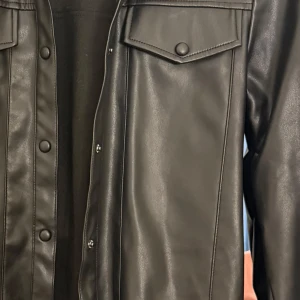 Svart skinnjacka från Pimkie - Snygg svart skinnjacka köpt ifrån nelly men märket heter Pimkie med knappar och bälte i midjan. Jackan har långa ärmar och en stilren design som passar perfekt för en cool look. Perfekt för dig som vill ha en edgy stil! Köpt för 600kr säljer den för 300