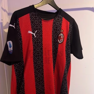 AC Milan fotbollströja från Puma - Snygg AC Milan fotbollströja från Puma i klassiska röd-svarta ränder. Tröjan har korta ärmar och klubbens emblem på bröstet. Perfekt för alla Milan-fans som vill visa sitt stöd på matchdagar! 🖤❤️