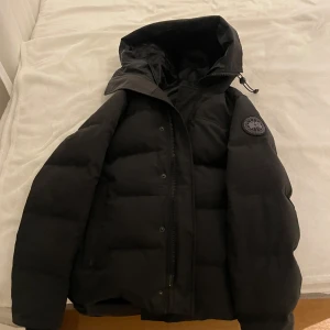 Svart dunjacka med huva - Säljer en svart dunjacka från Canada Goose med huva och dragkedja. Modellen heter Macmillan Parka black label. Jackan har en broderad logotyp på ärmen och är perfekt för kalla dagar. Den är långärmad och har en stilren design. Inköpt för drygt 3 månader sedan och allt kommer med. Kvitto, Box, hängare med mera. Nypris ligger på 14600. Fler bilder kan skickas vid önskemål. 