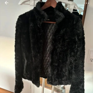 Svart pälsjacka - Snygg svart pälsjacka med quiltat foder. Jackan har en klassisk look med långa ärmar och en mjuk, fluffig yta. Perfekt för att hålla sig varm och stilren under kyliga dagar.
