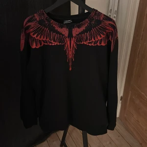 Svart tröja med röd vingdesign från Marcelo Burlon - Säljer en svart tröja från Marcelo Burlon med en cool röd vingdesign över axlarna och bröstet. Den är i storlek XL igentligen men har krympt till en L.
