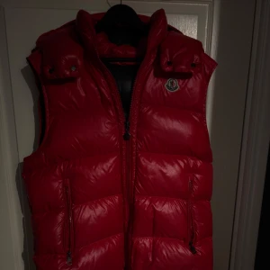 Moncler väst  - Snygg röd Moncler väst som såklart är äkta. Västen är storlek 4 som motsvarar L fast passar även M. Det finns ett jätte litet hål på framsidan av västen som man ser på 3 bilden. Hör av er för fler frågor! 
