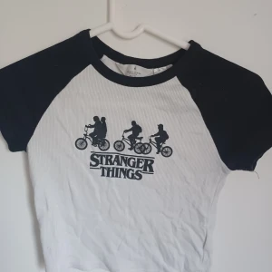 Vit och svart Stranger Things t-shirt - Säljer en cool vit och svart t-shirt med Stranger Things-tryck. T-shirten har korta ärmar och ett klassiskt raglanmönster. Perfekt för fans av serien!