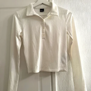 Vit ribbad långärmad tröja från Gina Tricot - Säljer en stilren vit ribbad tröja från Gina Tricot. Tröjan har långa ärmar och en klassisk krage med knappar framtill. Perfekt för en avslappnad men ändå chic look.