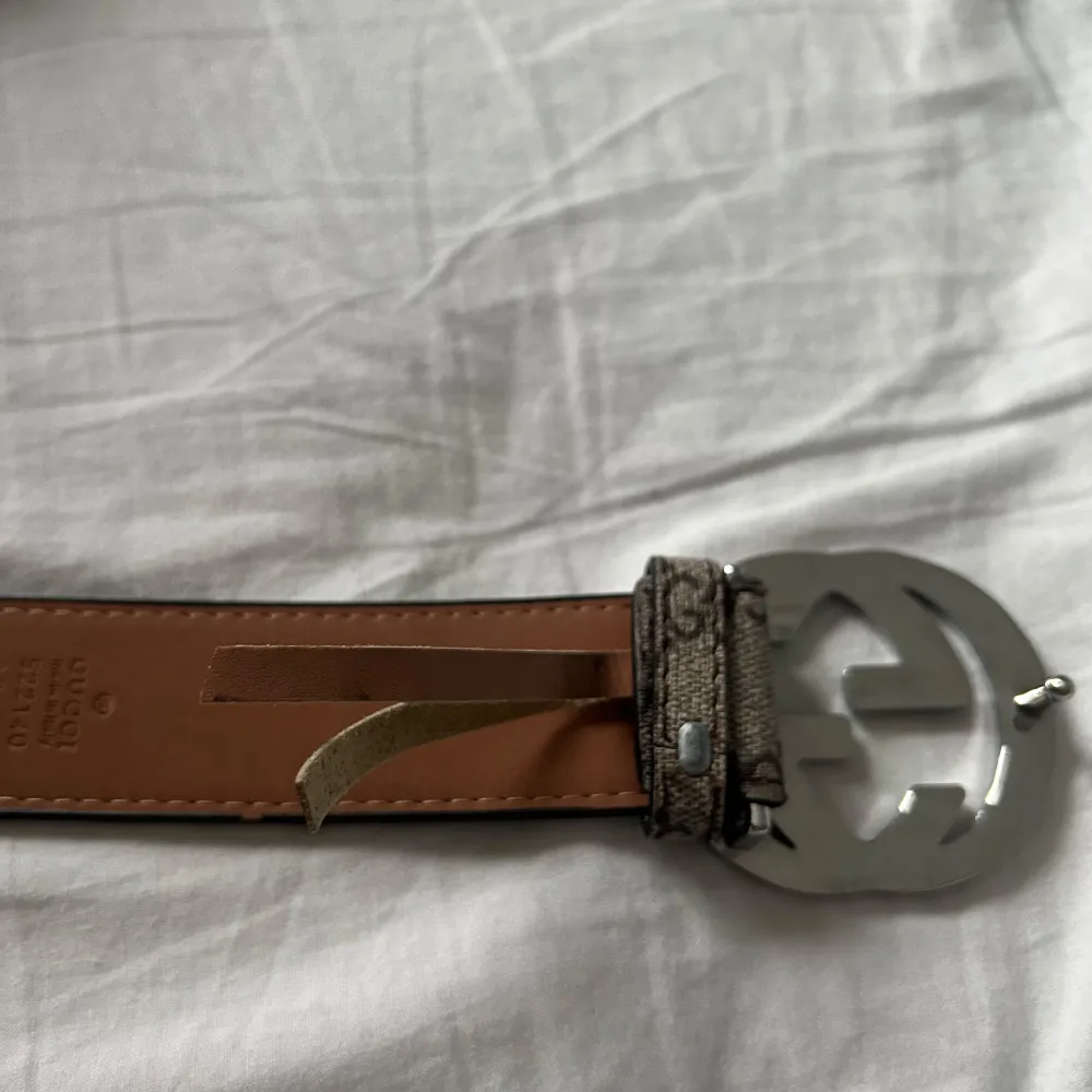Snyggt bälte från Gucci med ett klassiskt mönster och en stor, rund spänne i silver. Bältet har en stilren design med en brun insida och mönstrad utsida. Perfekt för att ge en lyxig touch till din outfit. Den är sönder lite men funkar jätte bra ändå. Man behöver inte ta bältet igenom första flärpen där den är sönder!. Asusteet.