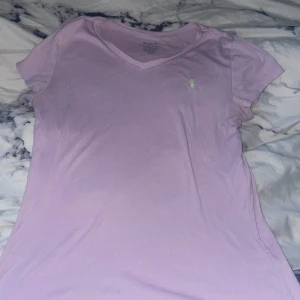 Ljuslila t-shirt från Polo Ralph Lauren - Säljer en ljuslila t-shirt från Polo Ralph Lauren med en grön broderad logga på bröstet. T-shirten har en v-ringad hals och korta ärmar. Det står storlek L men passar mig som har storlek Xs 