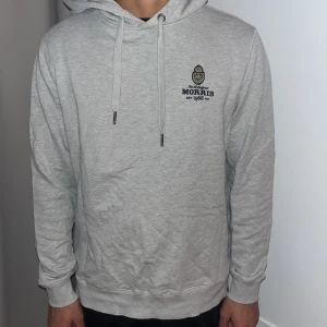 Grå hoodie från Morris - Säljer en stilren grå hoodie från Morris med broderad logga på bröstet. Tröjan har en klassisk passform med långa ärmar och justerbar huva med snören. Perfekt för en avslappnad stil.