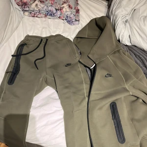 Olivgrön hoodie och byxor från Nike - Säljer en olivgrön hoodie och matchande byxor från Nike. Hoodien har dragkedja och byxorna har snörning i midjan. Båda plaggen har svarta detaljer och Nike-loggan på bröstet och benet. Perfekt för en avslappnad stil.