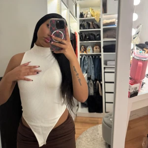 Vit ribbad ärmlös body - Snygg vit ribbad body med hög hals och ärmlös design. Perfekt för en stilren look. Toppen har en unik asymmetrisk nederdel som ger en modern touch.