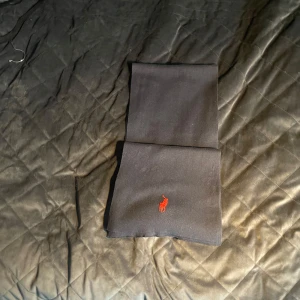 Svart halsduk från Polo Ralph Lauren - Snygg svart halsduk från Polo Ralph Lauren med en liten röd logotyp. Perfekt för att hålla sig varm och stilren under kyliga dagar. Halsduken är enkel och elegant, vilket gör den lätt att matcha med olika outfits.