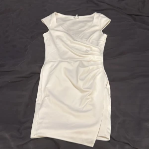 Vit klänning från NLY One - Elegant vit klänning från NLY One med omlott-design och v-ringning. Klänningen har en figurnära passform och är ärmlös, vilket ger en stilren look. Perfekt för studenten!