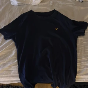 Mörkblå t-shirt från Lyle & Scott - Snygg mörkblå t-shirt från Lyle & Scott med deras ikoniska gula logga på bröstet. Perfekt för en stilren och avslappnad look. T-shirten har en klassisk passform och är kortärmad.