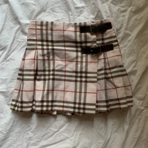 Rutig kjol från Burberry - Snygg rutig kjol från Burberry i ljusrosa och bruna toner. Kjolen har ett klassiskt rutmönster och två dekorativa spännen på sidan. Perfekt för en stilren look! Barnstorlek, passar XXS !!!🤩🤩🔥 klicka gärna på köp nu