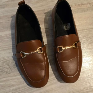 Bruna loafers med gulddetalj - Säljer ett par snygga bruna loafers i skinn med en elegant gulddetalj över foten. De har en klassisk design och ser riktigt stiliga ut. Perfekta för både vardag och fest! Står storlek 39 men är egentligen en 38
