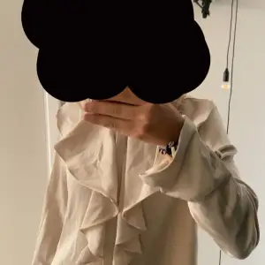 Säljer en elegant beige blus med vackra volangdetaljer framtill. Perfekt för både vardag och festliga tillfällen. Blusen har långa ärmar och en mjuk, bekväm passform.