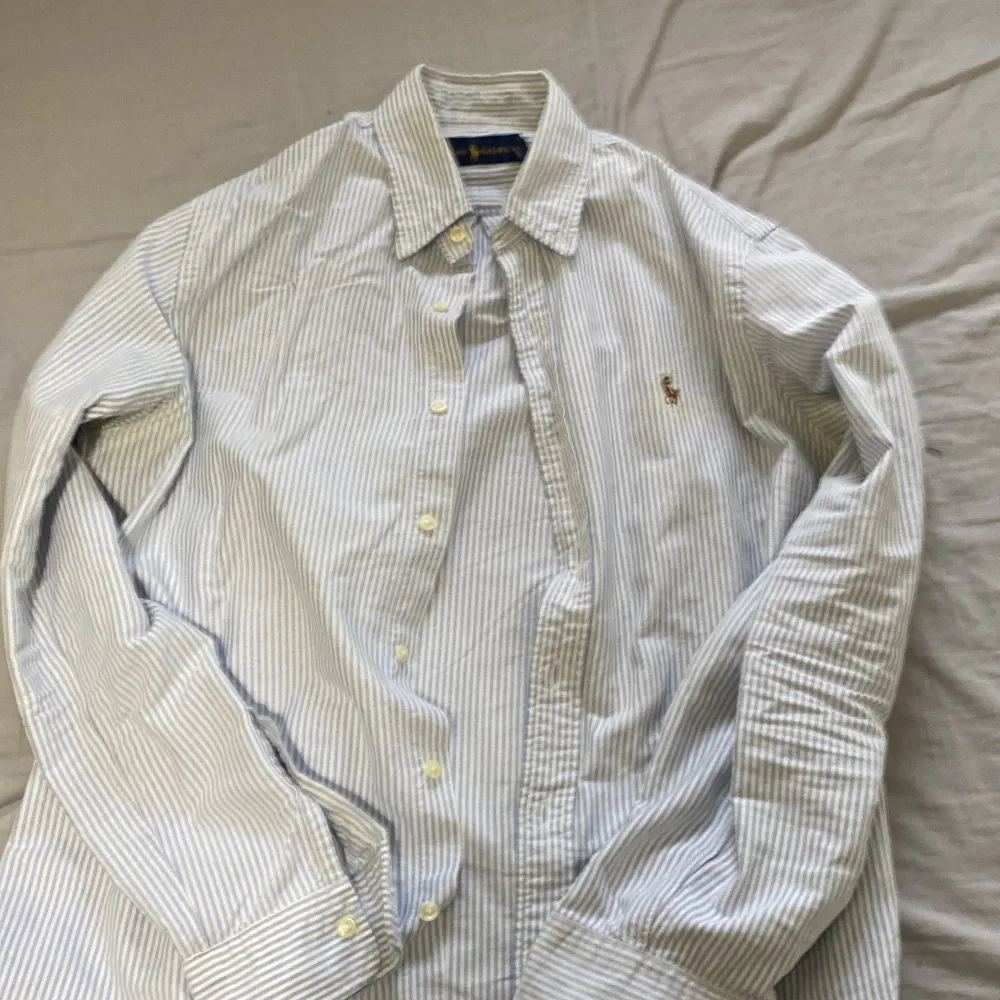 Säljer denna blåa Ralph lauren skjorta pga använder inte den så mycket och behöver pengarna. Den vita färgen håller på att fadea bort på några ställen tex vid under armarna och vid loggan och insidan, men det märker man inte av när man har på den.. Skjortor.