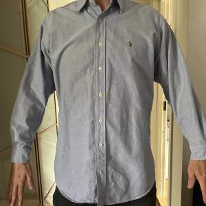 Blå skjorta från Ralph Lauren, storlek 16/33, vilket motsvarar typ storlek M. 16 tum är halsvidden och 33 tum är armlängden. Personen på bilden är 183 cm lång och väger ca 80 kg.