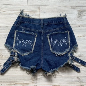 Jeansshorts med spännen 🔥  - Coola blåa ripped jeansshorts med justerbara spännen på sidorna och flames på bakfickorna 🔥Oanvända! 