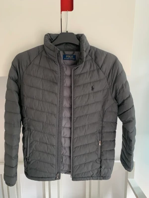 Grå dunjacka från Ralph lauren  - Grå tunn dunjacka från Ralph lauren köpt i usa typ 2018. En liten reva /hål i ena fickan men det går inte hela vägen in utan bara det yttre tyget. Inget dun åker ut och det är nog lätt att sy igen. Ganska sliten i helhet. Barnstorlek 