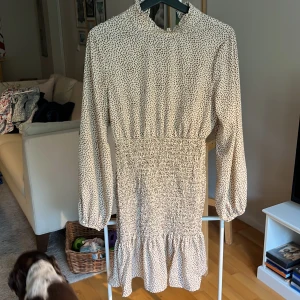 Söt klänning med volang - En användbar klänning då den är lika snygg med ett par sneakers till som klackskor. Beige med bruna små prickar (ser nästan ut som små hjärtan). Ballongärmar och volang nedtill. Som ny. Längd 90 cm. 