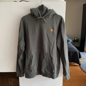 Hoodie Olivgrön - ”Organic” olivgrön Hoodie. Den har en slags muffgrej i luvan. Vill man inte ha den under hakan eller i ansiktet kan man bara trä huvudet framför.. annars går det att klippa av.  Hoodien är mer färgstark i verkligheten.