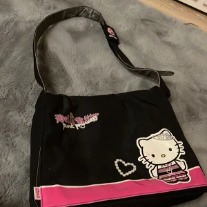 Hello Kitty väska - En hello Kitty väska med axelrem. Den är svart utanpå och rosa inuti. Väskan har jättegulliga hellokitty detaljer😺💕Skriv om ni vill ha flera bilder! 