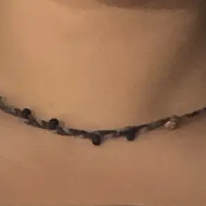 Ett flätat halsband med blå och brun tråd med pärlor och snäckor runt om. Den är handgjord i slitstarkt material med enkelt spänne som går att justera storleken på