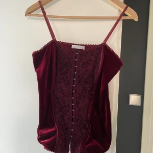Korsett  - Korsettliknande topp i vinröd färg. Står ingen storlek men uppskattar till S/M åt det större hållet. Plastskenor och knäpps framtill. I använt men gott skick.