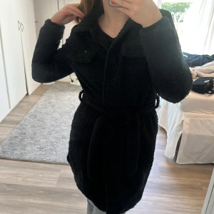 Vår jacka - En svart lite tjockare vår jacka, som är fluffig och med bruna knappar på vänster sida.  Medellång i längden. 