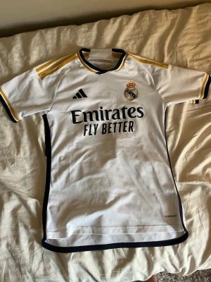 Real Madrid matchtröja Vini Jr 7 - Säljer en vit Real Madrid matchtröja från Adidas med svarta och gula detaljer. Tryck med Vini Jr och nummer 7 på ryggen, klubbmärke och sponsorlogga på bröstet. Materialet är lätt och andas, perfekt för fotbollsträning eller match.