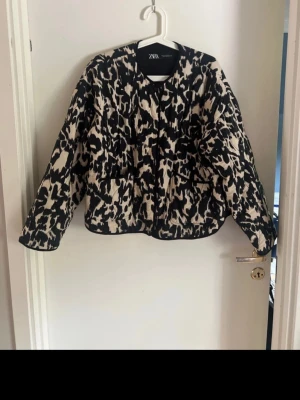 Leopardmönstrad quiltad jacka Zara - Snygg quiltad jacka från Zara med leopardmönster i svart och beige. Jackan har rund halsringning, knappar framtill och en loose passform. Perfekt för dig som vill sticka ut med ett trendigt mönster.