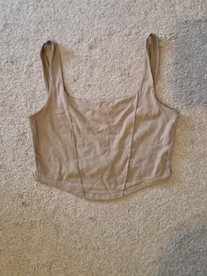 Beige korsett topp  - Säljer en enkel och stilren beige croppad linnetopp i korsett stil, med breda axelband och rundad halsringning. Toppen har en figurnära passform.