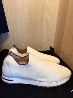Loro Piana Sneakers - Säljer ett par stilrena vita slip-on sneakers med stickad ovandel och bruna detaljer runt öppningen. Skorna har en vit sula med diskreta ränder och är perfekta för dig som gillar en clean och modern look. Materialet är mjukt och flexibelt för extra komfort.