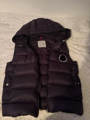 Svart dunväst med huva från Moncler - Snygg svart dunväst från Moncler med huva och quiltad design. Västen har två fickor med tryckknappar, dragkedja framtill och en dekorativ Moncler-logga på bröstet. Perfekt för lager-på-lager och streetwear-stil.