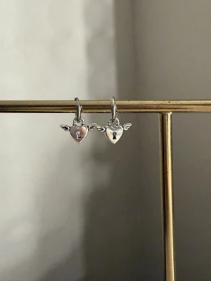 Silverörhängen hjärta med vingar - Säljer ett par söta örhängen i silverfärg med hjärtformade hängen och små vingar på sidorna. Hängena har ett litet nyckelhål i mitten för extra detalj. Perfekta för dig som gillar unika och lekfulla smycken.