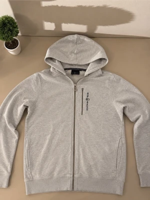 Ljusgrå hoodie från Sail Racing - Snygg ljusgrå hoodie från Sail Racing med dragkedja framtill och broderad logga på bröstet. Hoodien har två fickor på sidorna och en klassisk huva. Tillverkad i mjukt bomullsmaterial som känns skönt mot huden.