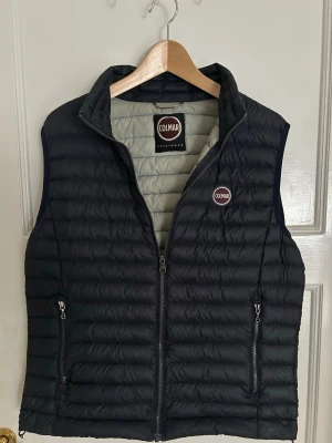 Mörkblå dunväst från Colmar storlek 52/L - Snygg mörkblå dunväst från Colmar med quiltad design och två dragkedjeförsedda fickor. Västen har hög krage och Colmar-logga på bröstet. Perfekt att slänga över en tröja för extra värme. Lätt och smidig att bära. En lagning men syns inte då kragen täcker den bra. En dragskedja på fickan är lite trög